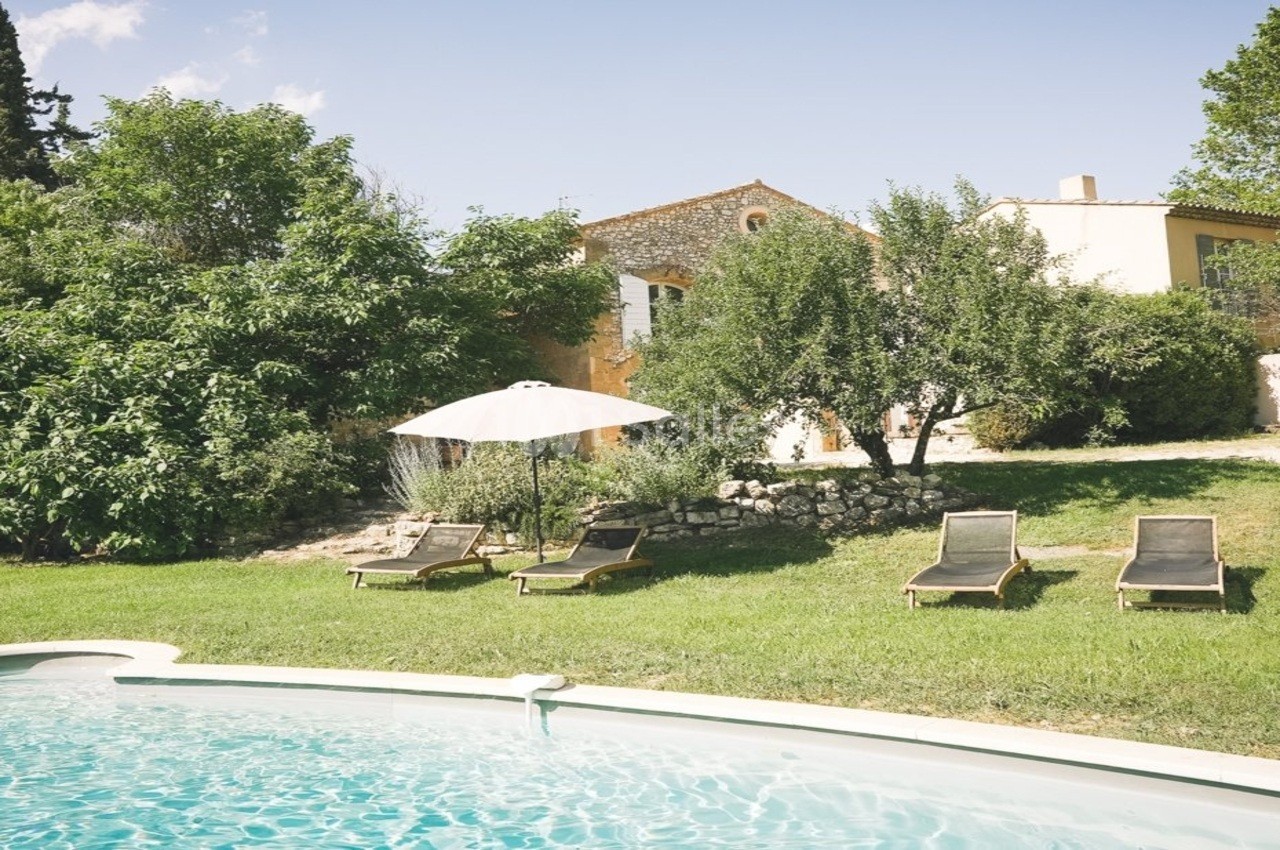 Jardin avec piscine, transats et parasol, entouré de verdure et d'une maison en pierre sous un ciel dégagé.
