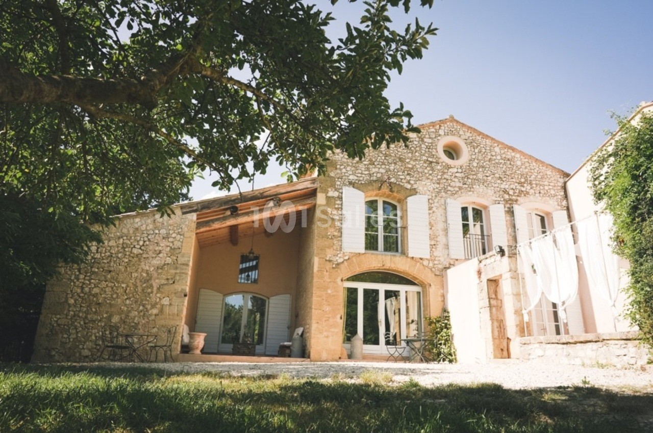 Façade en pierre d'une maison avec volets blancs, grande ouverture vitrée et terrasse ombragée par un arbre.