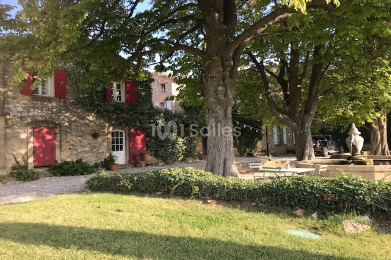 Façade en pierre d'une maison avec volets rouges, entourée d'arbres et d'un jardin verdoyant.