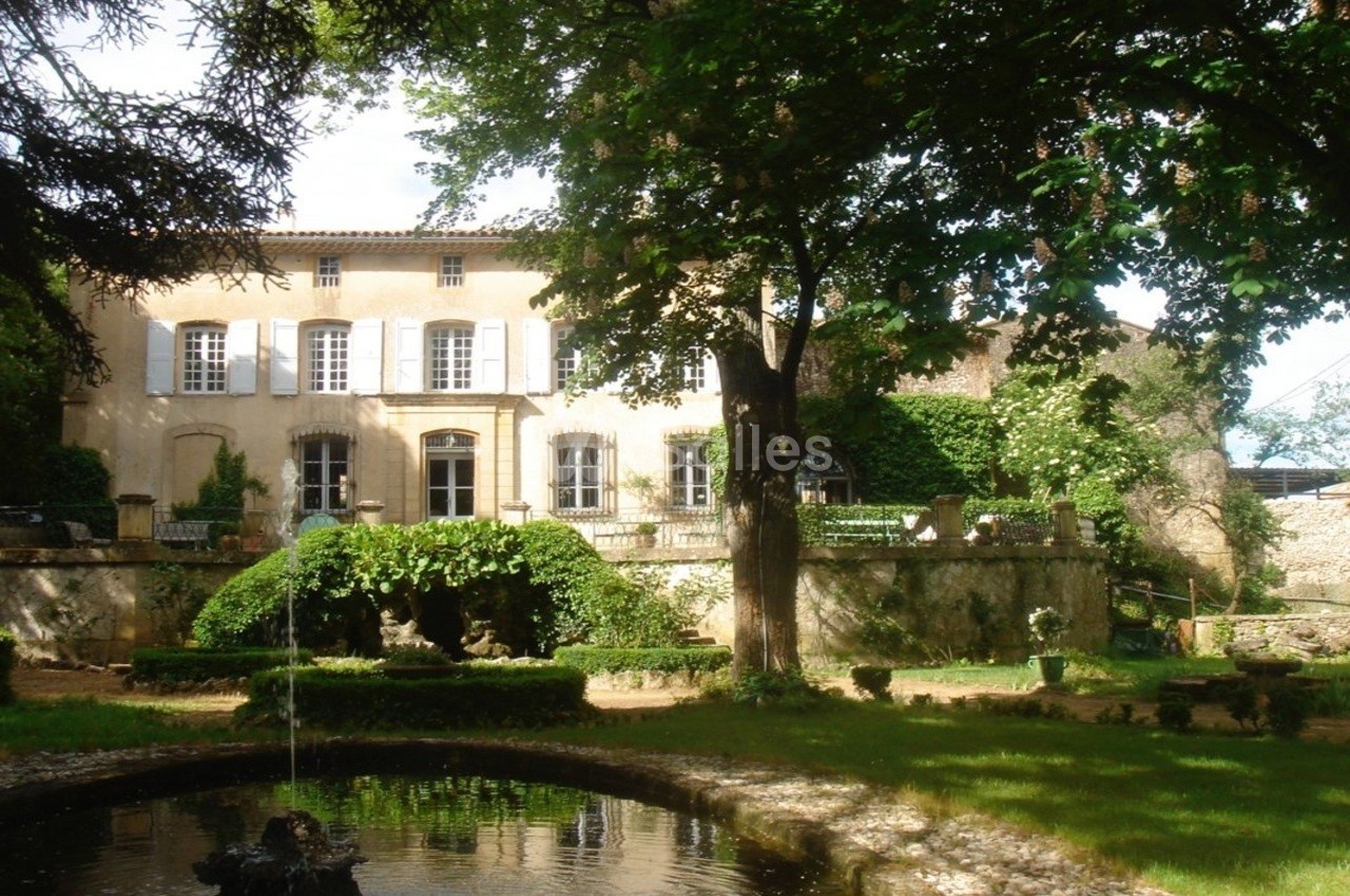 Façade d'une maison ancienne entourée de verdure, avec un jardin et une fontaine au premier plan.