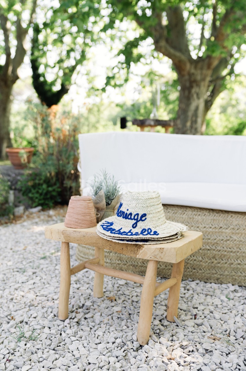 Chapeau en paille brodé posé sur un tabouret en bois, entouré de plantes et d'un canapé extérieur sous des arbres.