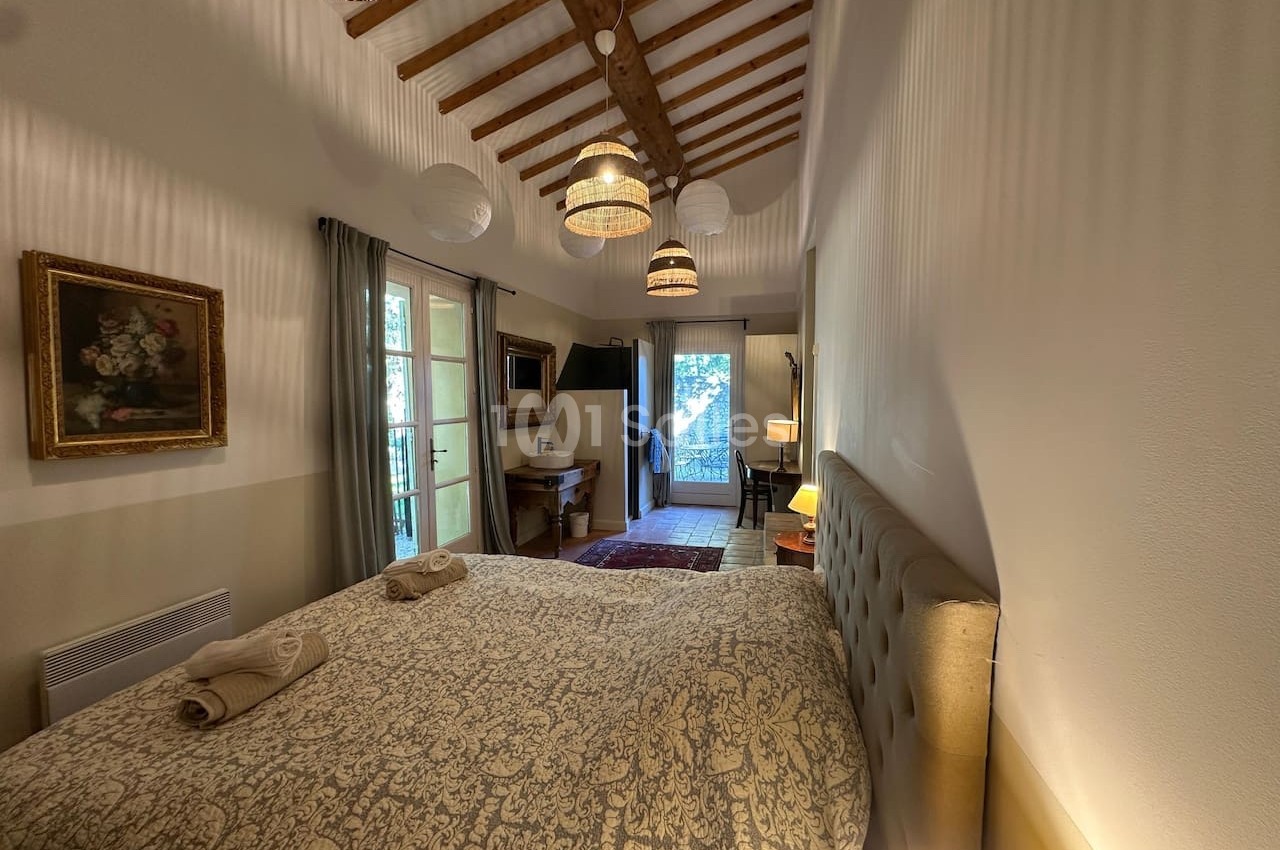 Chambre lumineuse avec lit double, poutres apparentes, décoration classique et vue sur un balcon arboré.