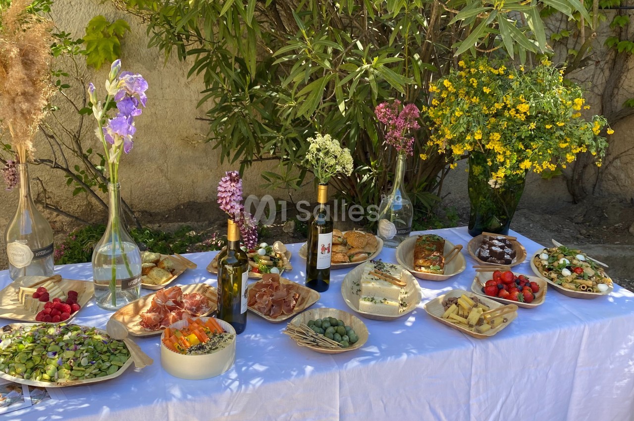 Table dressée en extérieur avec des plats variés, des fleurs en vase, des bouteilles de vin et un décor végétal en arrière…