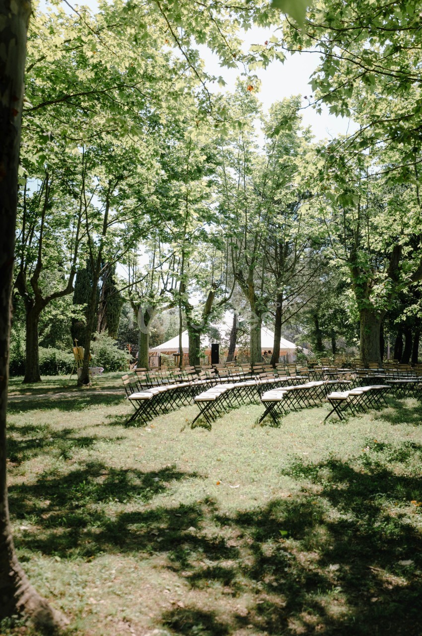 Chaises disposées en arc sous des arbres dans un parc, avec une tente blanche visible en arrière-plan.