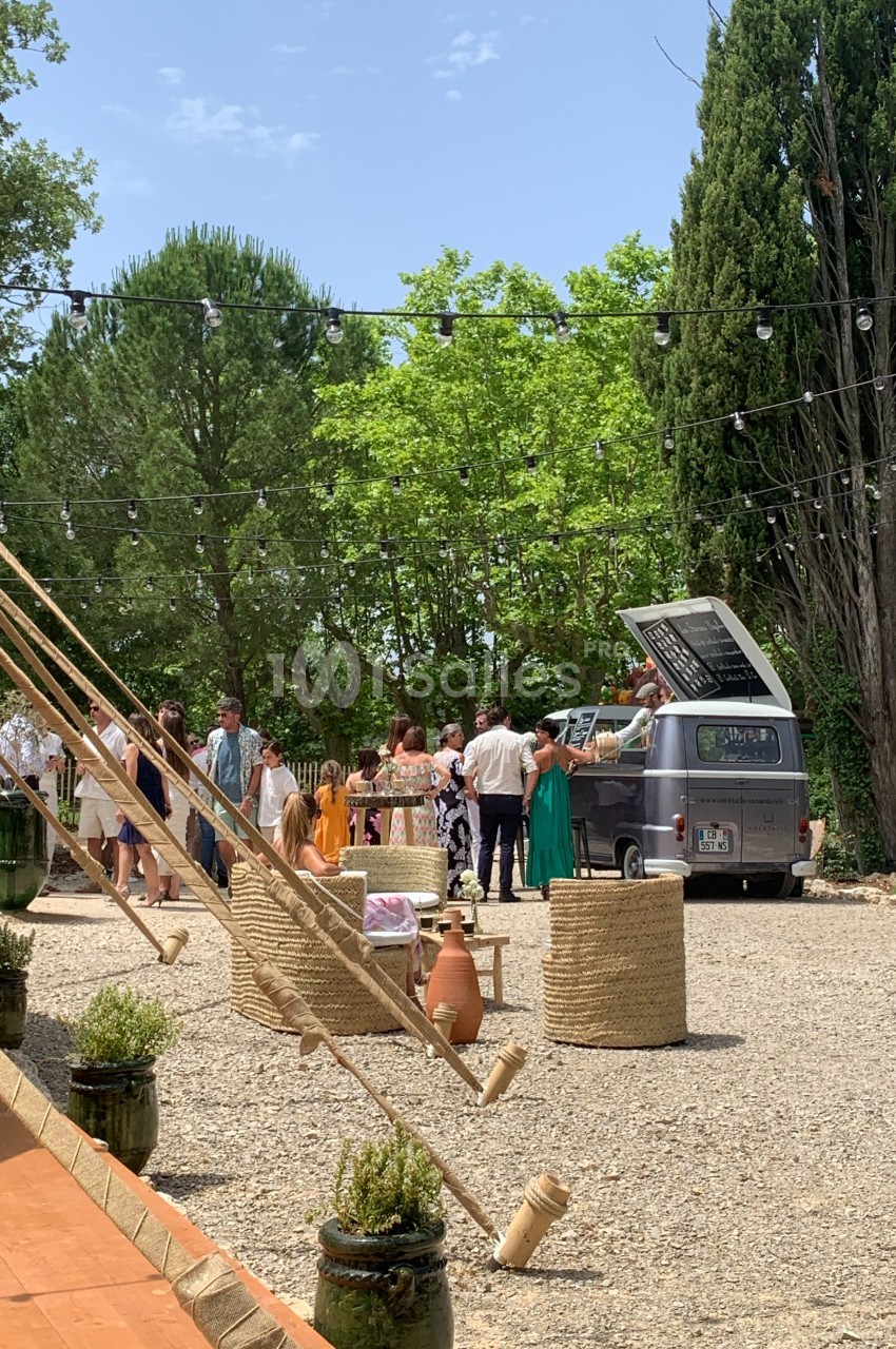 Un groupe de personnes rassemblé autour d'un camion-bar vintage dans un espace extérieur arboré et ensoleillé.