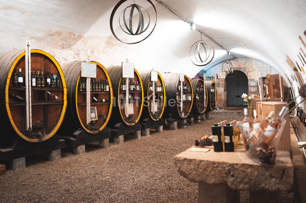 Cave voûtée avec des tonneaux en bois contenant des bouteilles de vin, et une table avec des produits locaux au premier plan.