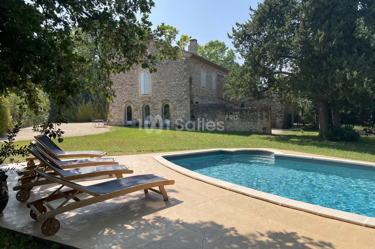 Maison en pierre entourée de verdure avec une piscine et des chaises longues en bois sur une terrasse ensoleillée.