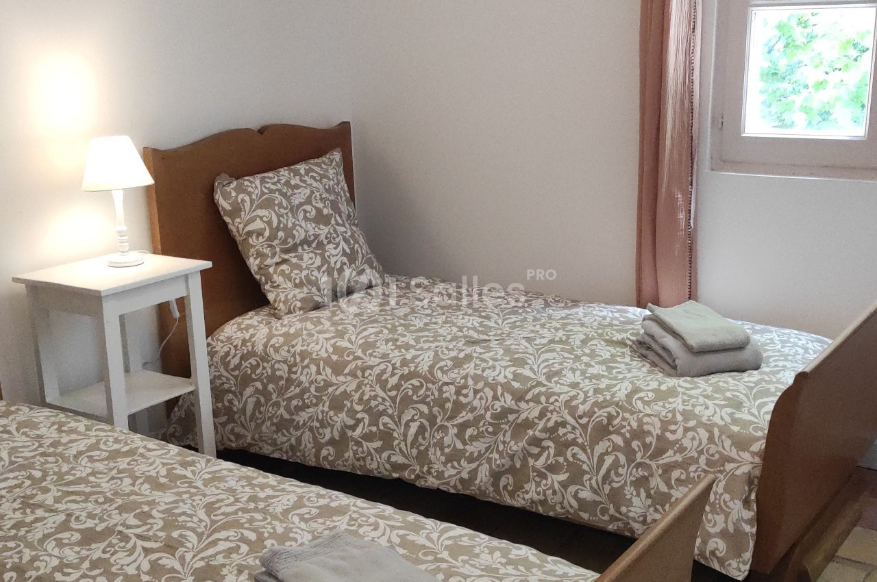 Deux lits simples avec linge de lit à motifs, une table de chevet avec lampe et une fenêtre laissant entrer la lumière.