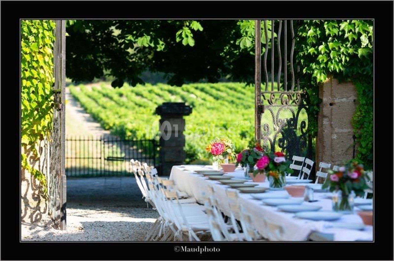 Table dressée en extérieur sous des arbres, avec vue sur un vignoble à travers une porte en fer forgé.
