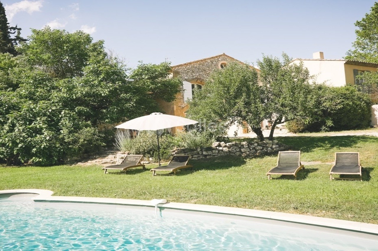 Jardin avec piscine, transats sous un parasol blanc et maison en pierre entourée de végétation.
