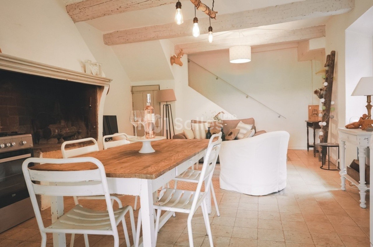Salle à manger rustique avec table en bois, chaises blanches, cheminée ancienne et salon en arrière-plan.