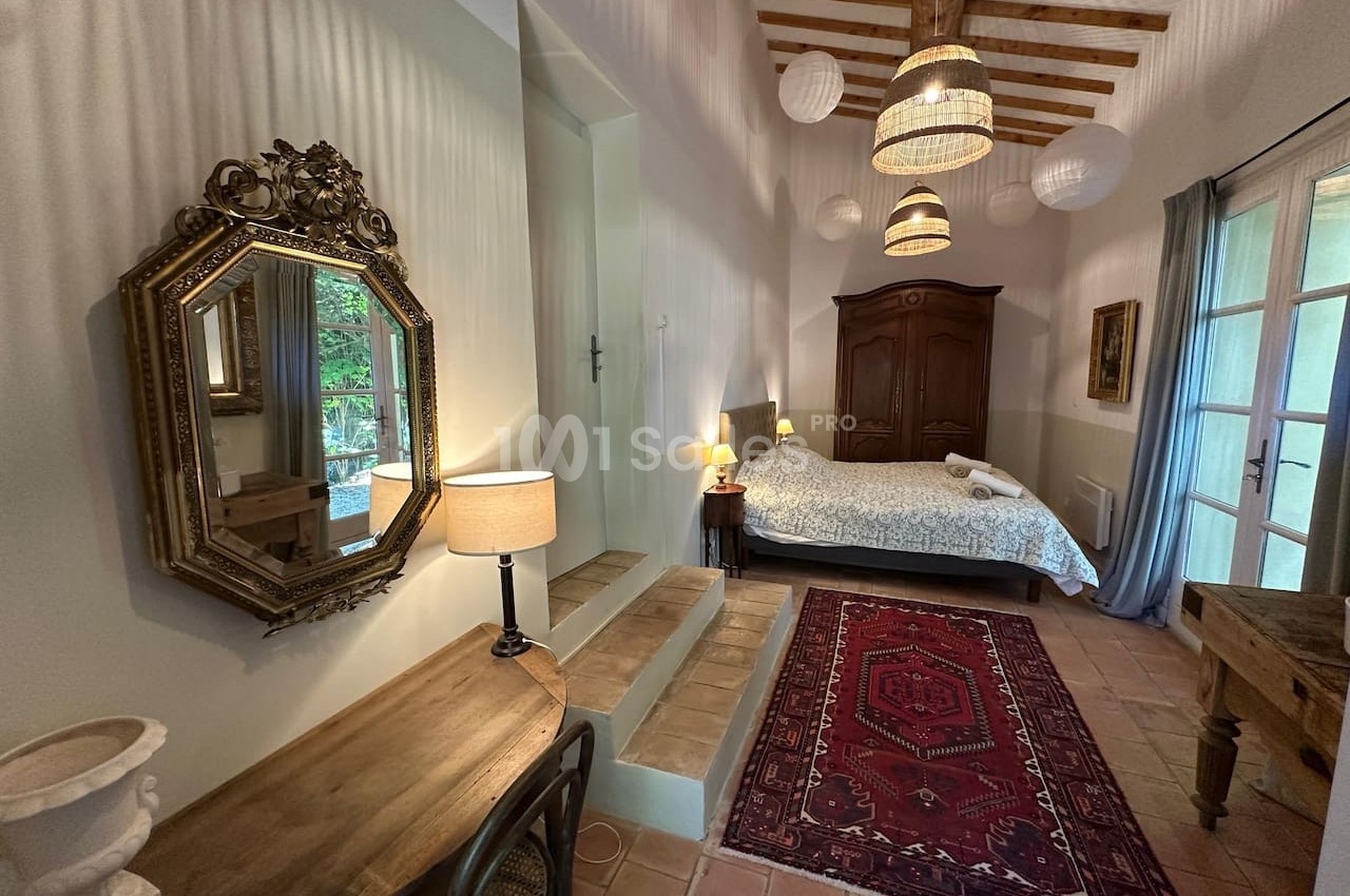 Chambre lumineuse avec lit double, bureau en bois, miroir orné, tapis rouge et plafond avec poutres apparentes.