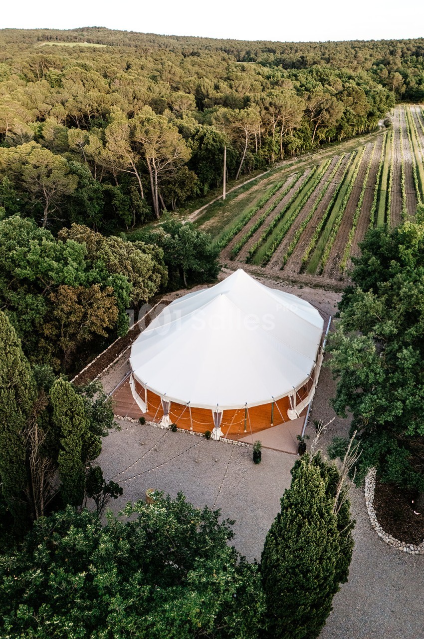 Chapiteau blanc et orange en plein air entouré de vignes et de forêt, vue aérienne.
