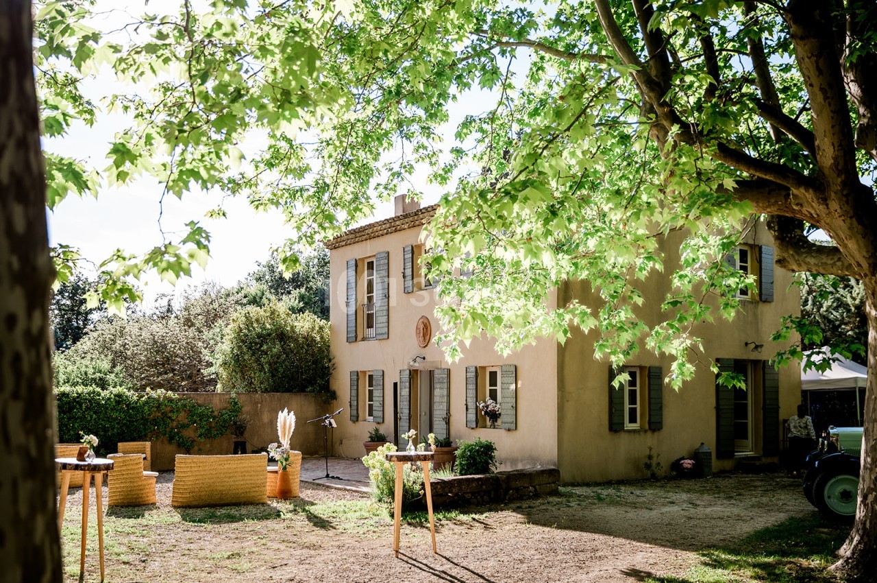 Maison provençale ocre à deux étages avec volets bleus, cour de gravier ombragée par un grand arbre feuillu.
