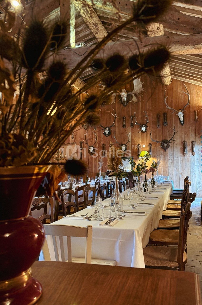 Salle rustique avec tables dressées, chaises en bois, trophées de chasse au mur et décoration florale.