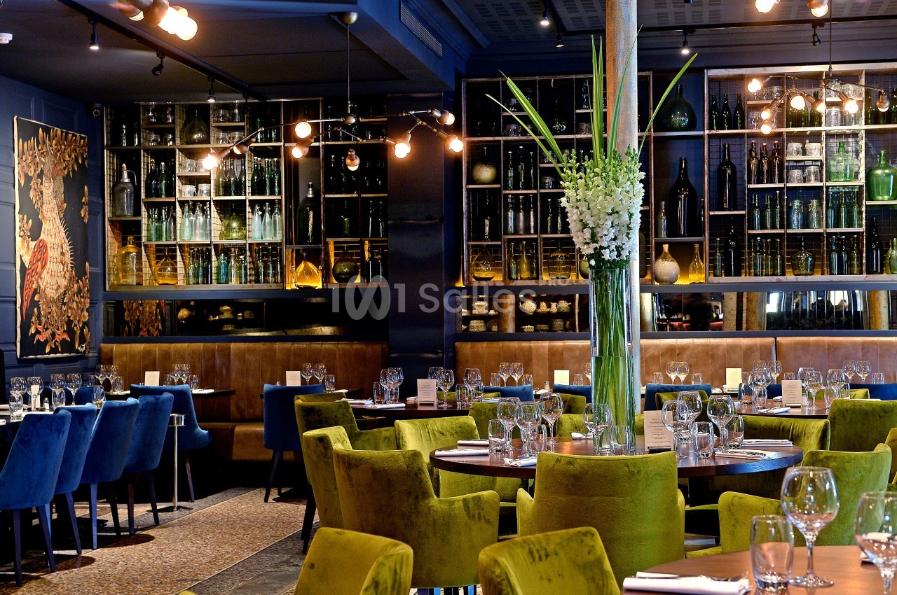 Salle de restaurant élégante avec des tables dressées, des chaises colorées et des étagères décorées de bouteilles et objets.