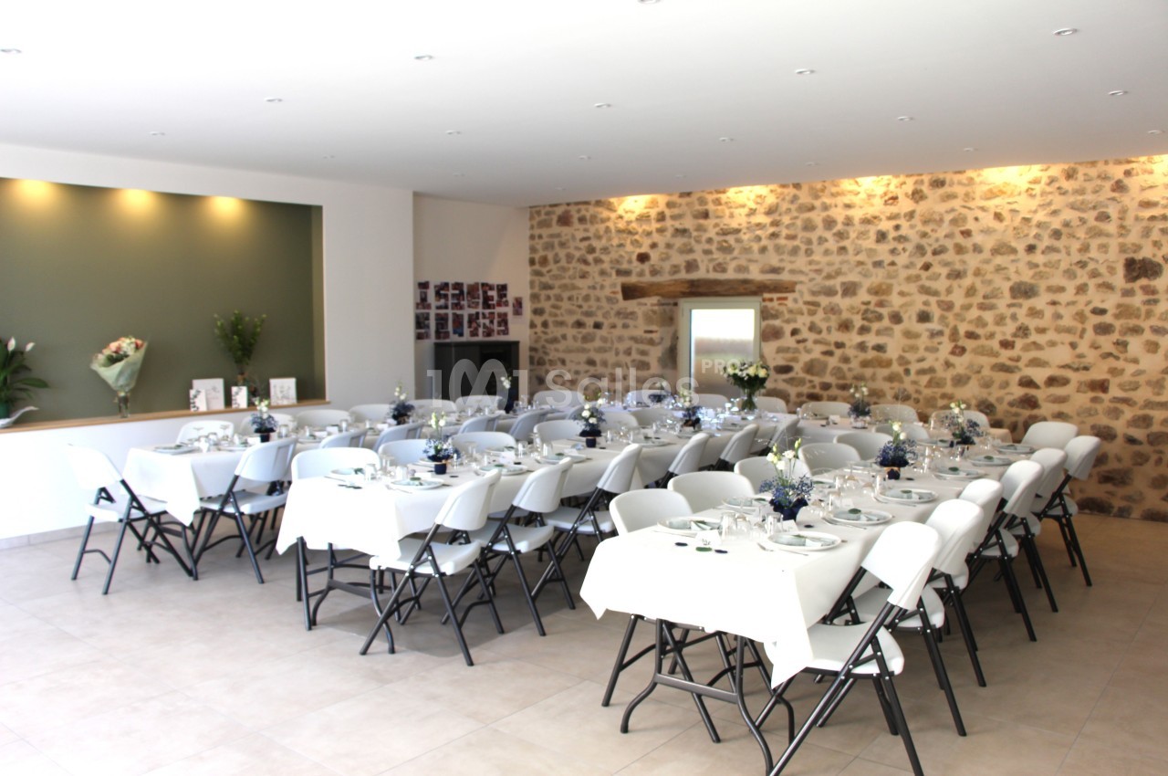 Salle de réception lumineuse avec des tables dressées, chaises blanches et murs en pierre apparente.