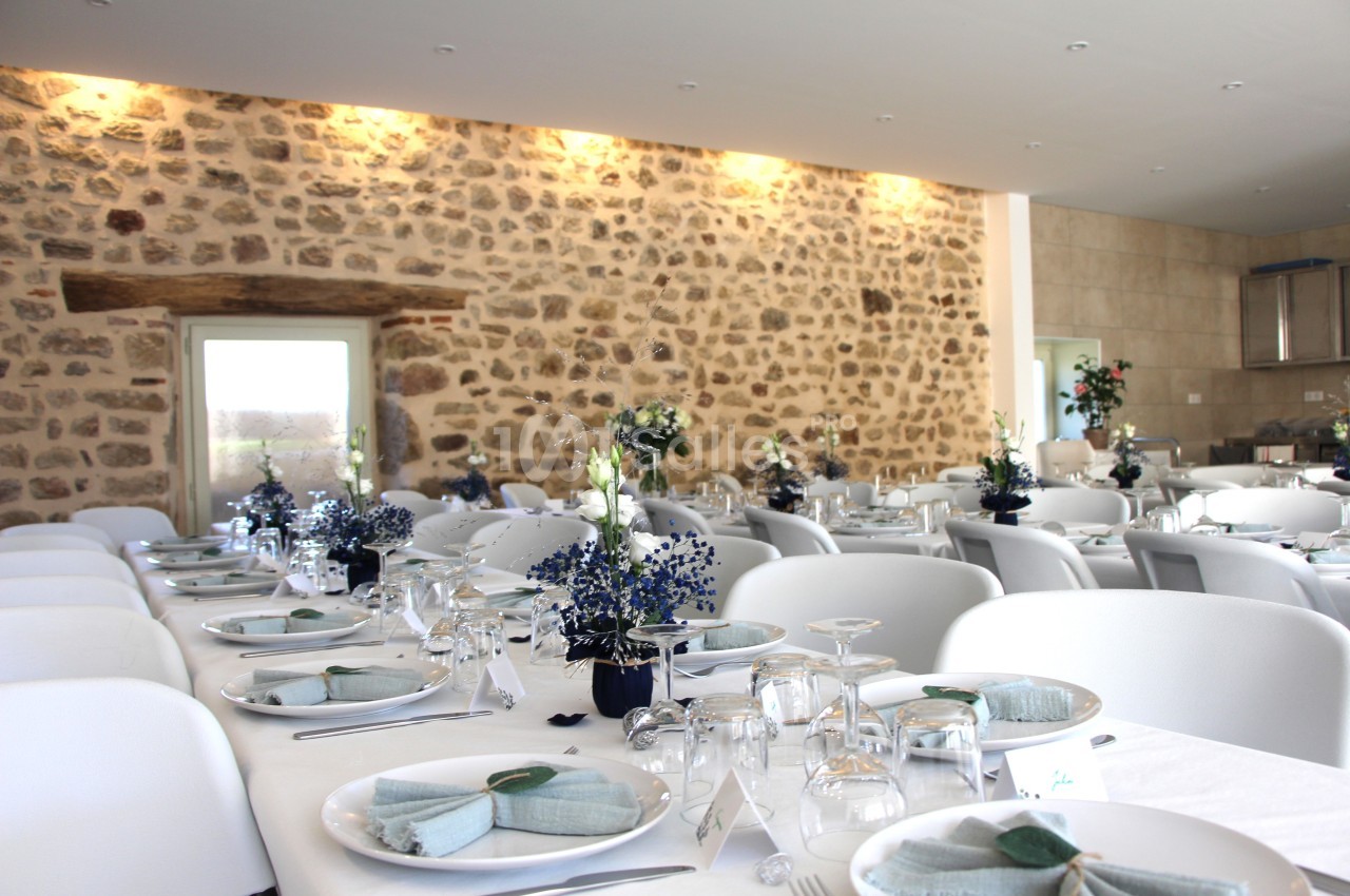 Salle de réception avec murs en pierre, tables dressées avec nappes blanches, vaisselle élégante et décorations florales.