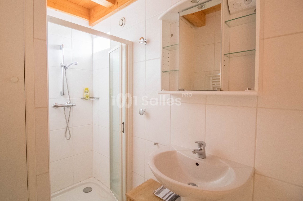 Salle de bain lumineuse avec douche à paroi vitrée, lavabo blanc, miroir avec étagères et plafond en bois.