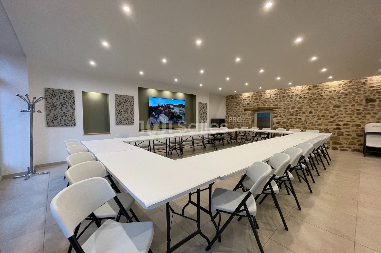 Salle de réunion lumineuse avec tables blanches disposées en U, chaises pliantes et mur en pierre apparente.