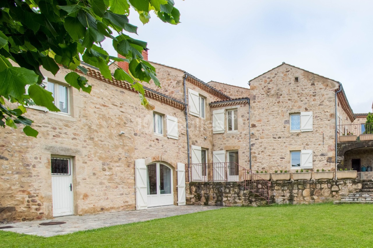 Façade en pierre d'une maison traditionnelle avec volets blancs, entourée d'une pelouse et d'arbres.