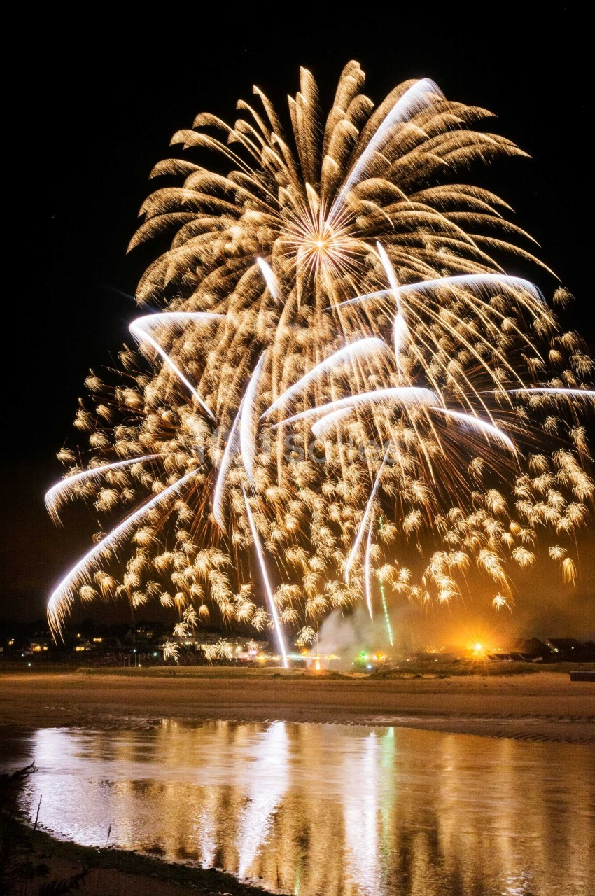 Feu d'artifice illuminant le ciel nocturne au-dessus d'une rivière, avec des reflets lumineux sur l'eau.