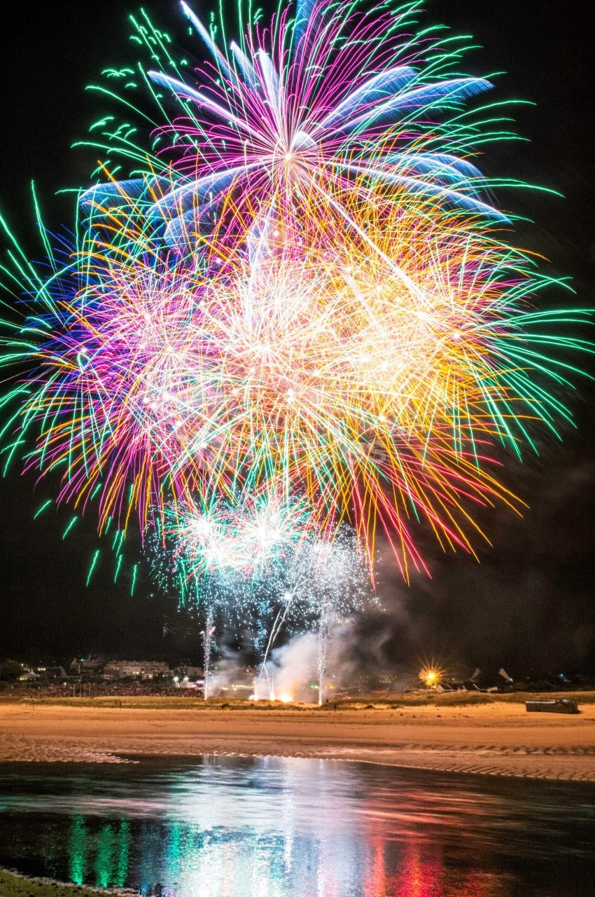 Feux d'artifice multicolores illuminant le ciel nocturne au-dessus d'une plage avec leur reflet dans l'eau.