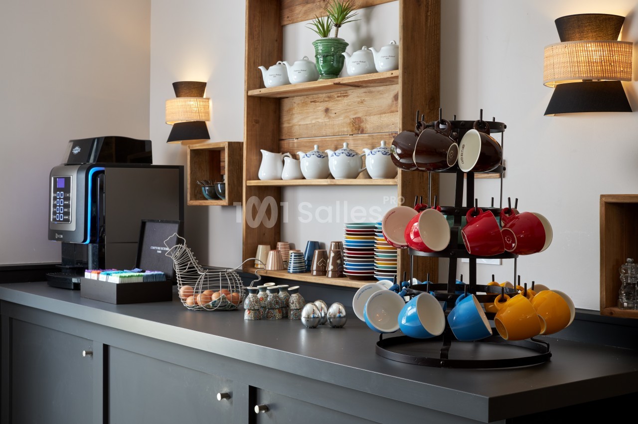 Comptoir avec machine à café, tasses colorées, pots en céramique et étagères décorées d'objets assortis.