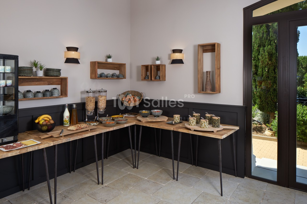Buffet de petit-déjeuner avec céréales, fruits, pain et boissons, disposé sur des tables en bois dans une salle lumineuse.