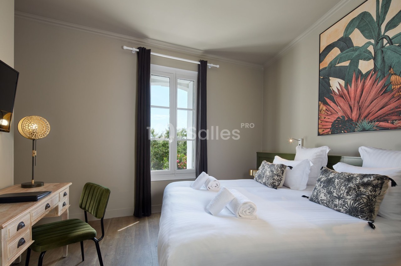 Chambre lumineuse avec lit double, bureau en bois, chaise verte, grande fenêtre et décoration murale colorée.