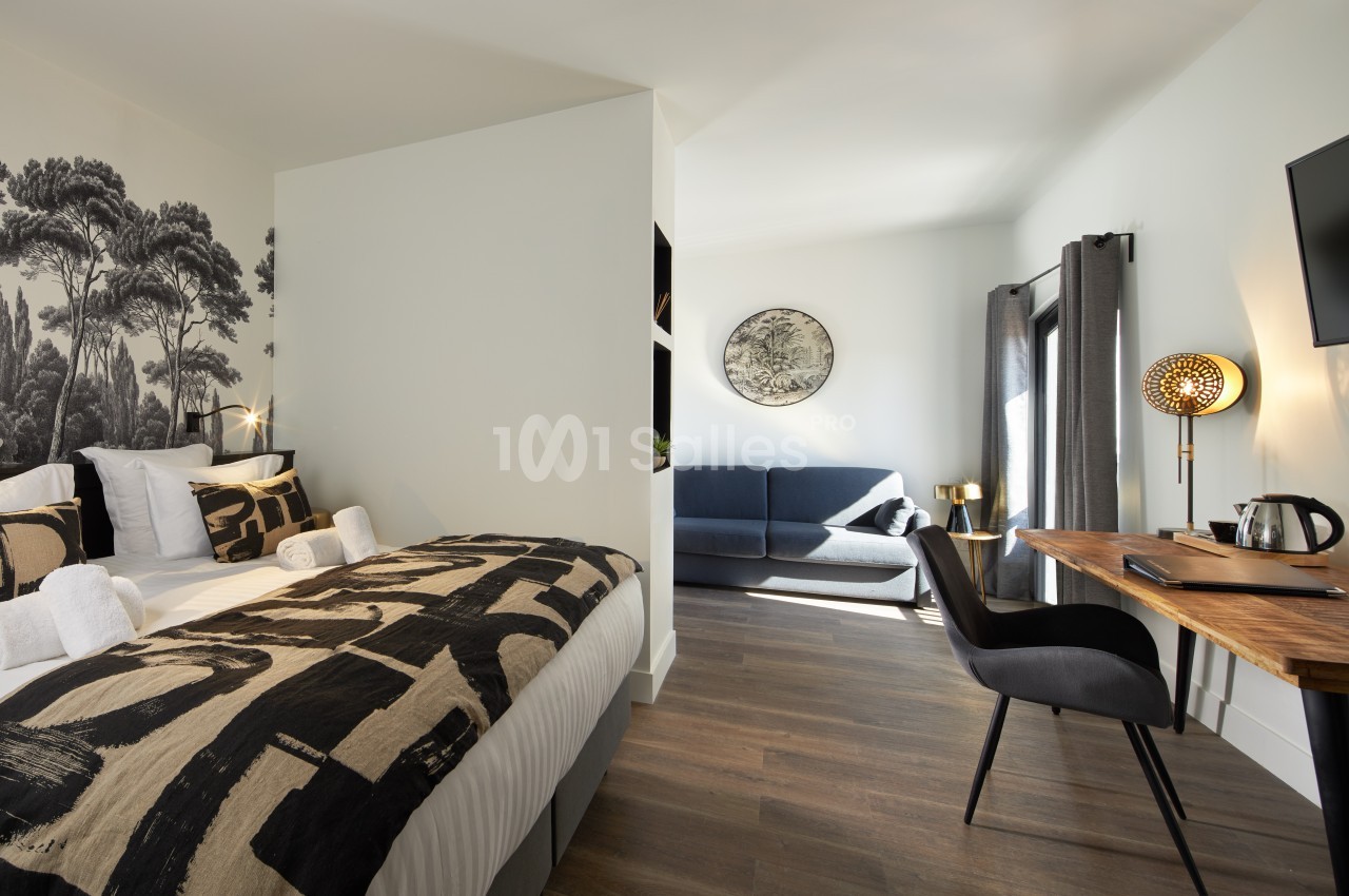 Chambre moderne avec lit double, bureau, canapé, décor mural et lumière naturelle entrant par une fenêtre.