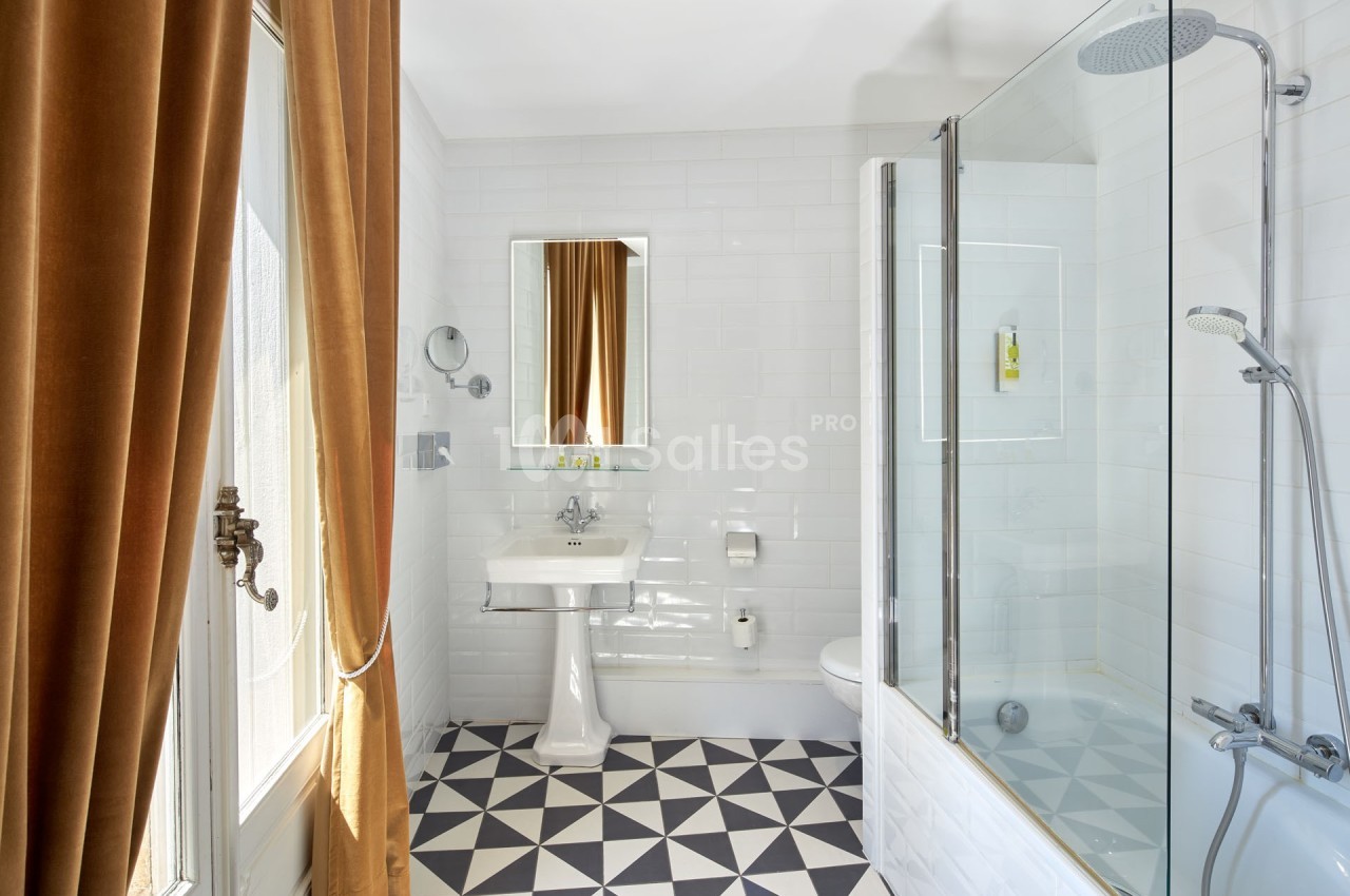 Salle de bain lumineuse avec carrelage géométrique, baignoire avec paroi vitrée, lavabo et rideaux marron.