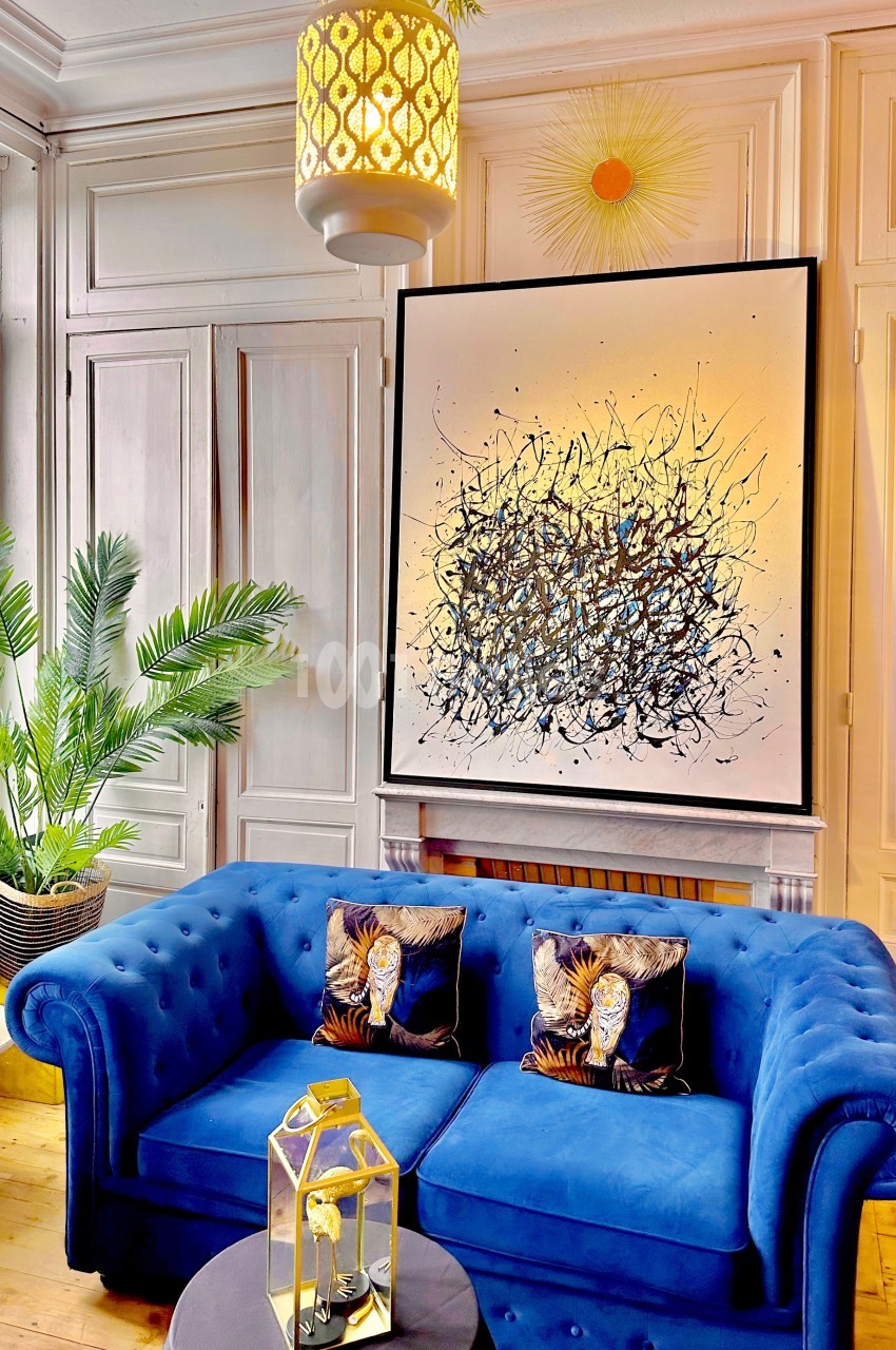 Canapé bleu avec coussins décoratifs, tableau abstrait au mur, plante verte et lampe suspendue dans un salon lumineux.