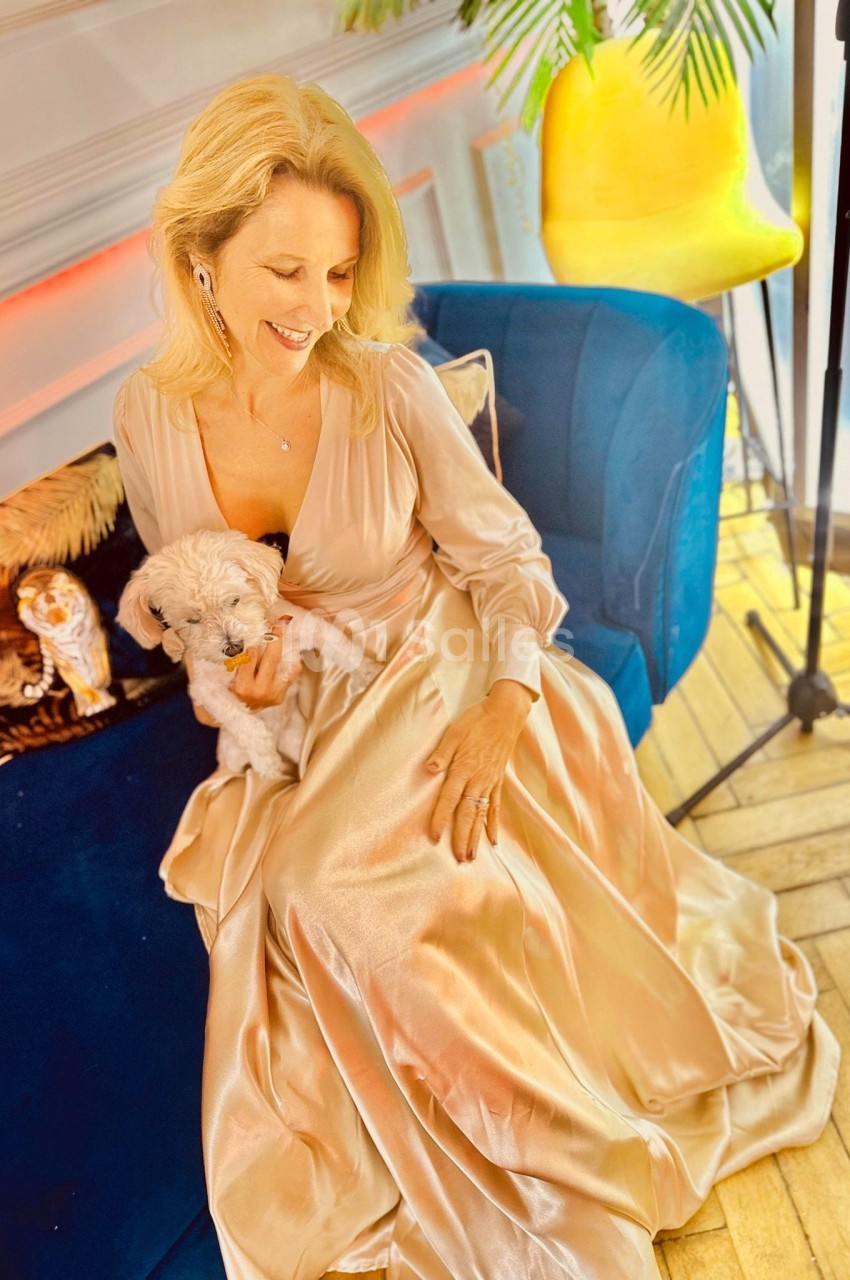 Femme souriante en robe satinée beige assise sur un canapé bleu, tenant un petit chien blanc sur ses genoux.