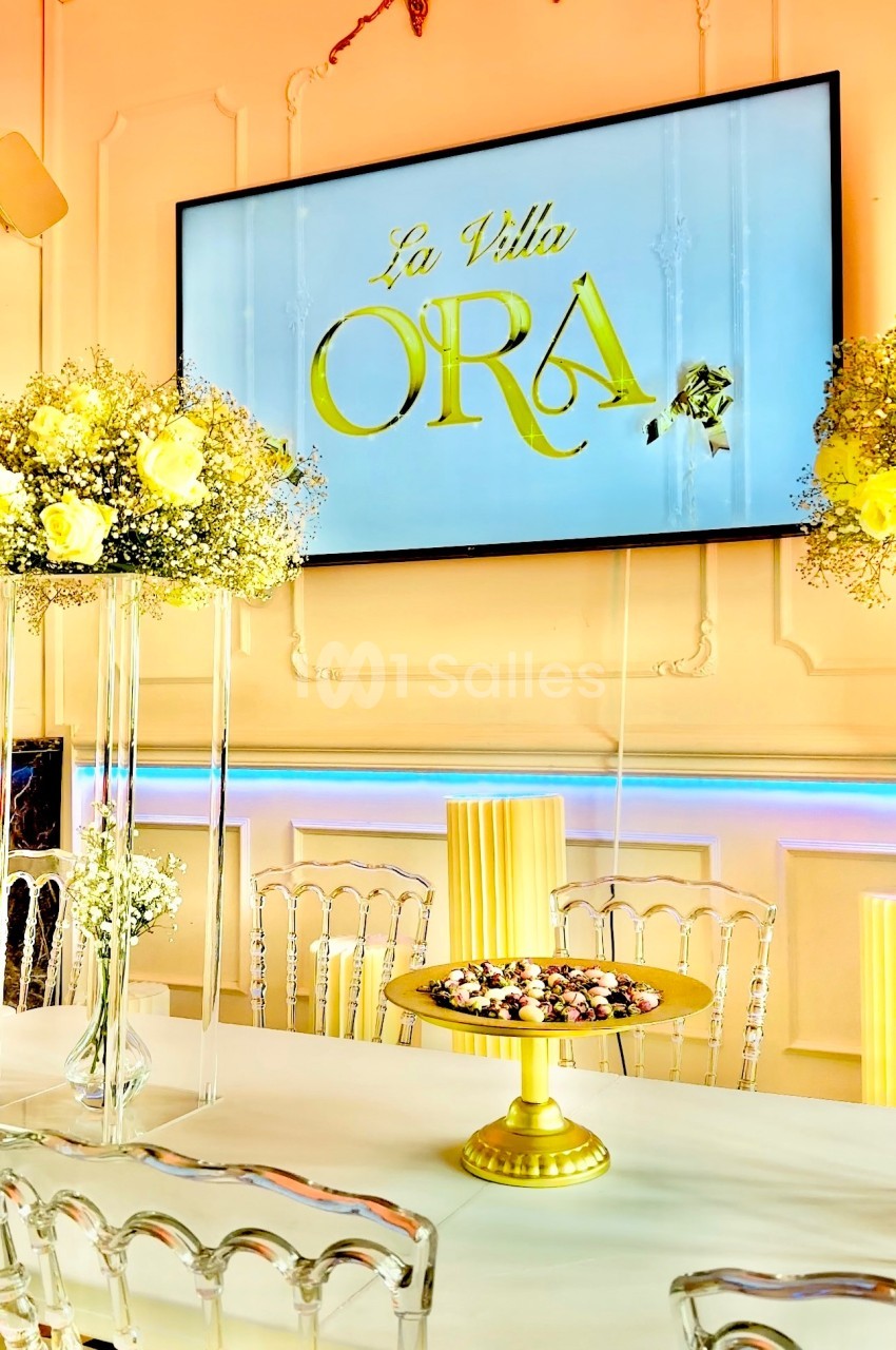 Salle décorée avec des chaises transparentes, une table ornée de fleurs et un écran affichant ’La Villa Ora’.