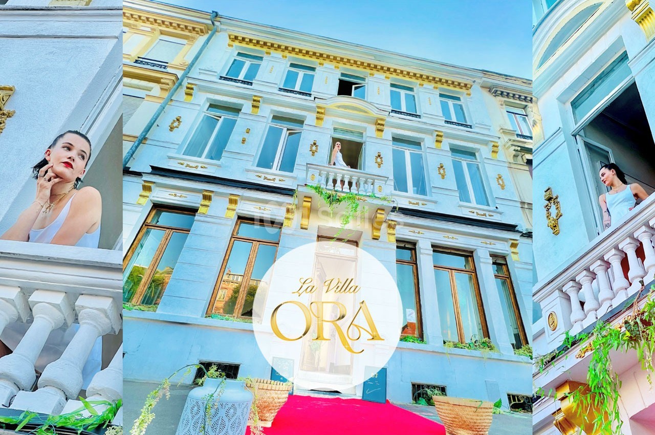 Façade d'une villa élégante avec balcons ornés, une femme en robe blanche visible sur un balcon.