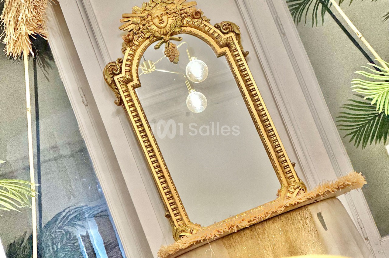 Miroir doré orné de motifs sculptés, posé devant une fenêtre avec des plantes en arrière-plan.