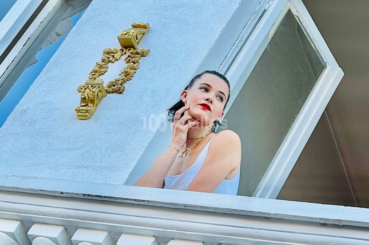 Femme en débardeur blanc regardant par une fenêtre ouverte, appuyée sur le rebord, avec un heurtoir doré visible.