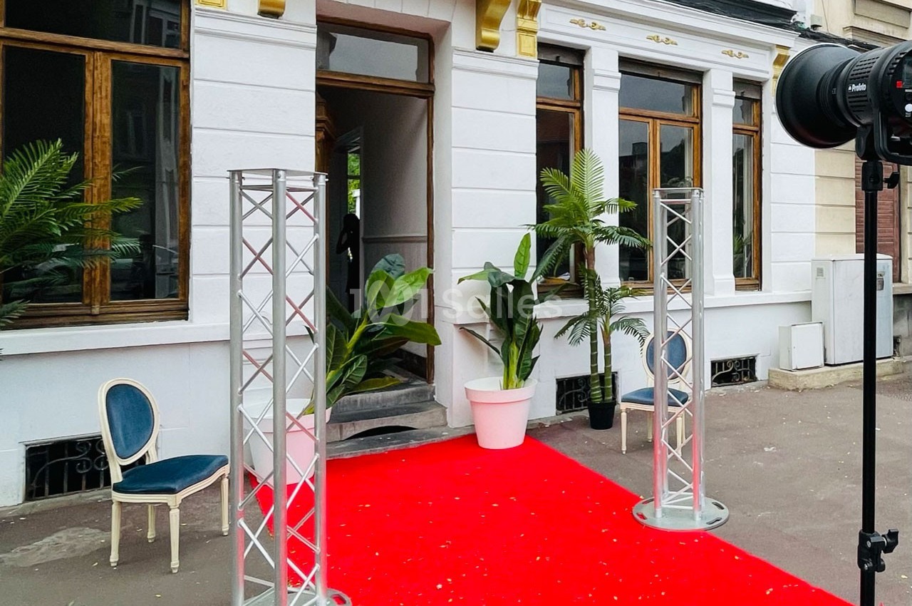 Entrée d'un bâtiment avec tapis rouge, plantes en pot, structures métalliques et chaise sur le côté.