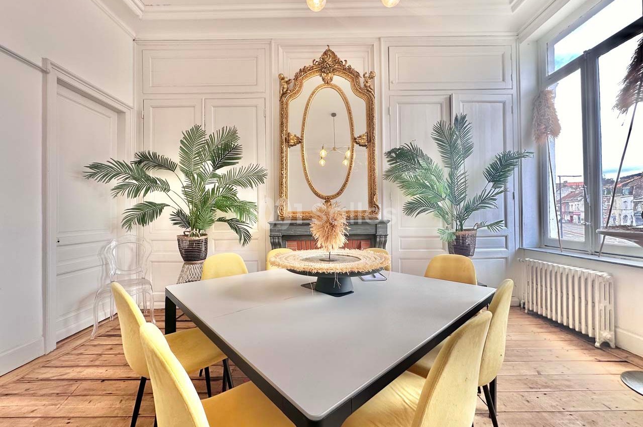 Salle à manger lumineuse avec table carrée, chaises jaunes, miroir doré, plantes et parquet en bois clair.