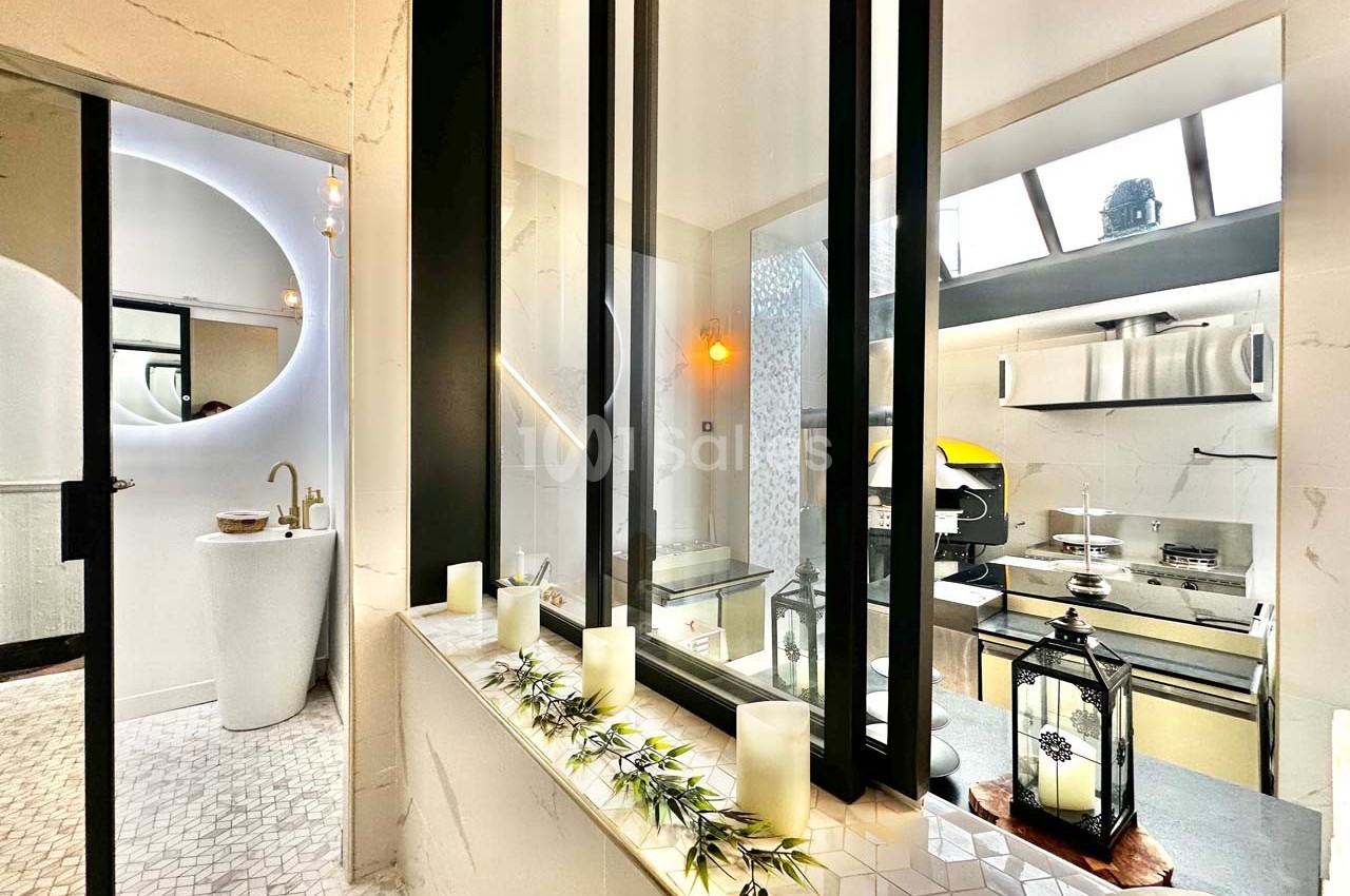 Salle de bain moderne avec lavabo design, bougies décoratives sur rebord et vue sur une cuisine extérieure lumineuse.