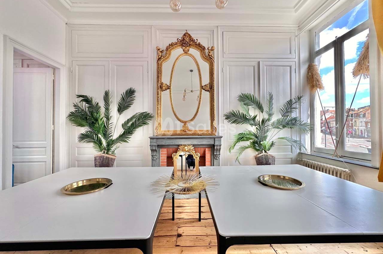 Salle lumineuse avec murs blancs, miroir doré, plantes décoratives, deux tables blanches et vue sur l'extérieur.