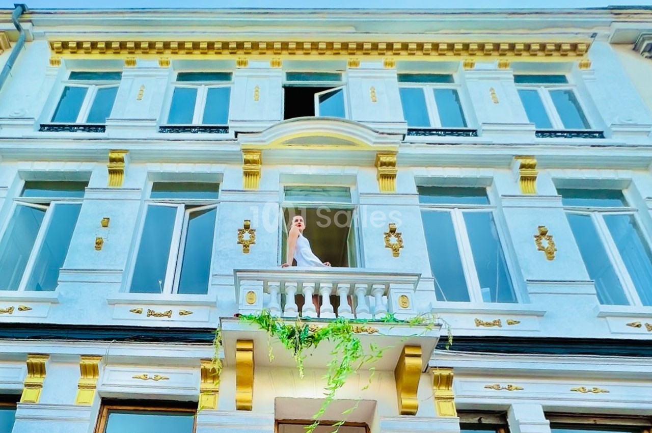 Femme en robe blanche debout sur un balcon orné de plantes, devant une façade élégante aux détails dorés.