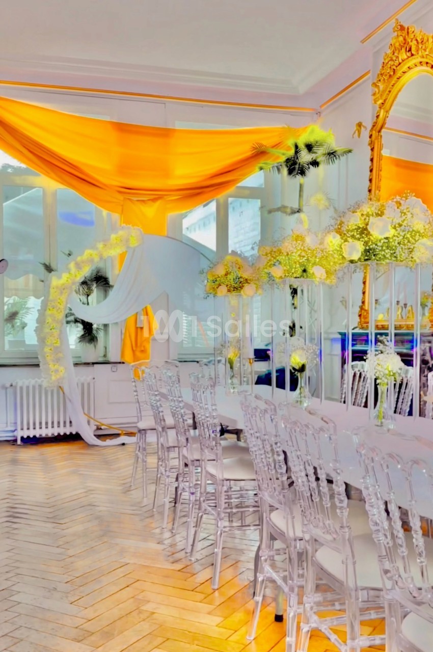 Salle décorée avec des chaises transparentes, une table blanche, des fleurs blanches et jaunes, et un drapé orange vif.