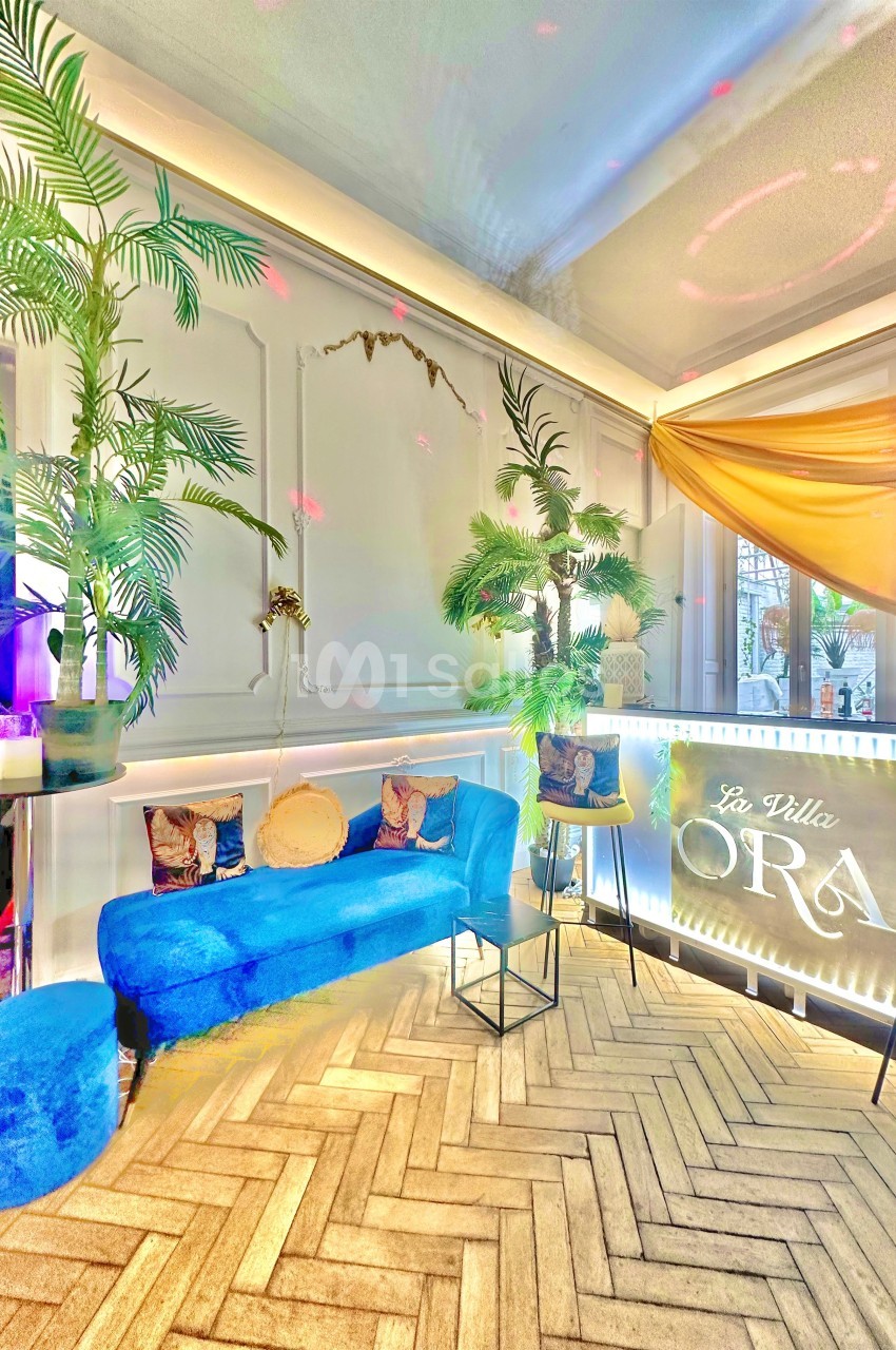 Salon lumineux avec canapé bleu, plantes tropicales, parquet en chevrons et enseigne lumineuse ’La Villa Ora’.