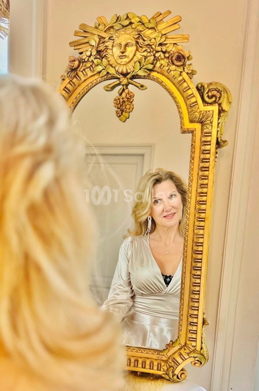 Femme blonde regardant son reflet dans un miroir doré orné de motifs sculptés.