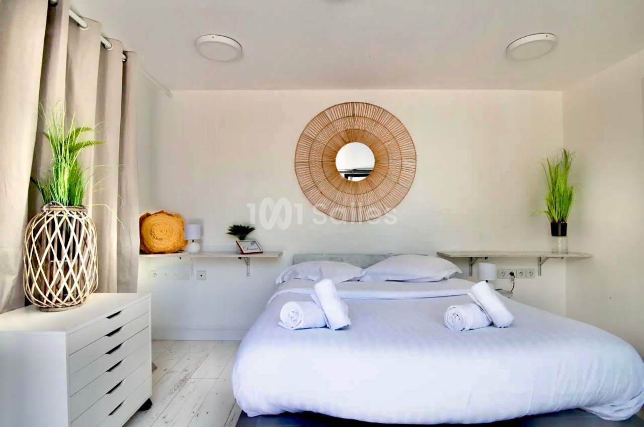 Chambre lumineuse avec lit double, miroir rond en rotin, plantes décoratives et mobilier blanc minimaliste.