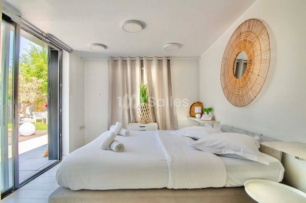 Chambre lumineuse avec lit double, décoration épurée, miroir rond en rotin et vue sur un jardin par une baie vitrée.