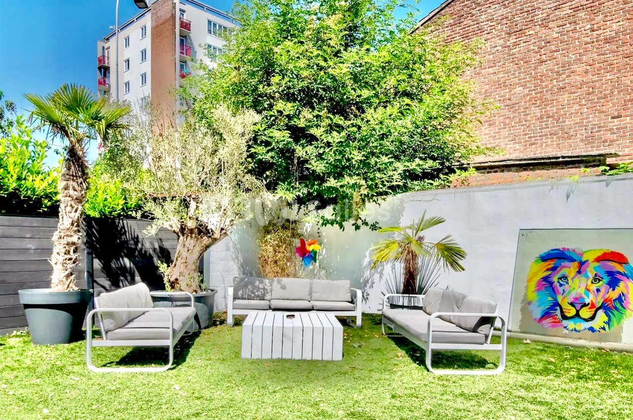 Salon de jardin avec canapés gris, table basse blanche, plantes exotiques et tableau coloré sur un mur extérieur.