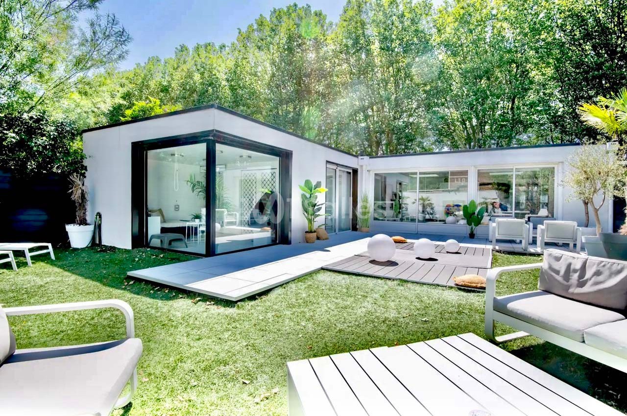 Maison moderne avec grandes baies vitrées, terrasse en bois, mobilier d'extérieur et jardin arboré en arrière-plan.